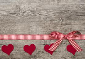 Red checkered ribbon with bow and four red felt hearts on a rustic wooden background.